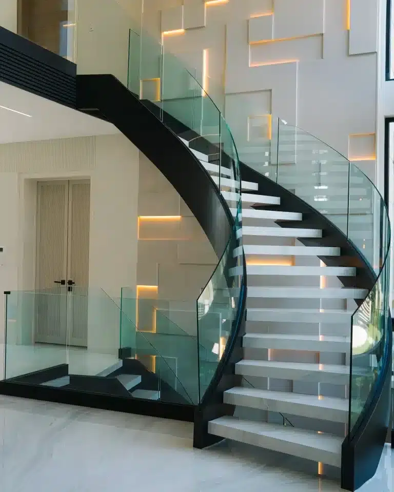 Curved Frameless Glass Railing Curved frameless glass railing in Toronto on a black steel staircase with white stone treads and a lit feature wall