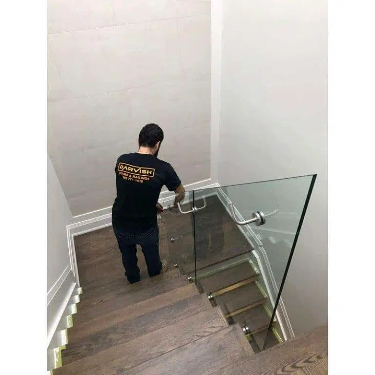 Darvish Crew During Glass Railing Installation Darvish Inc installer fitting a stainless steel handrail to a frameless glass railing on a dark oak staircase