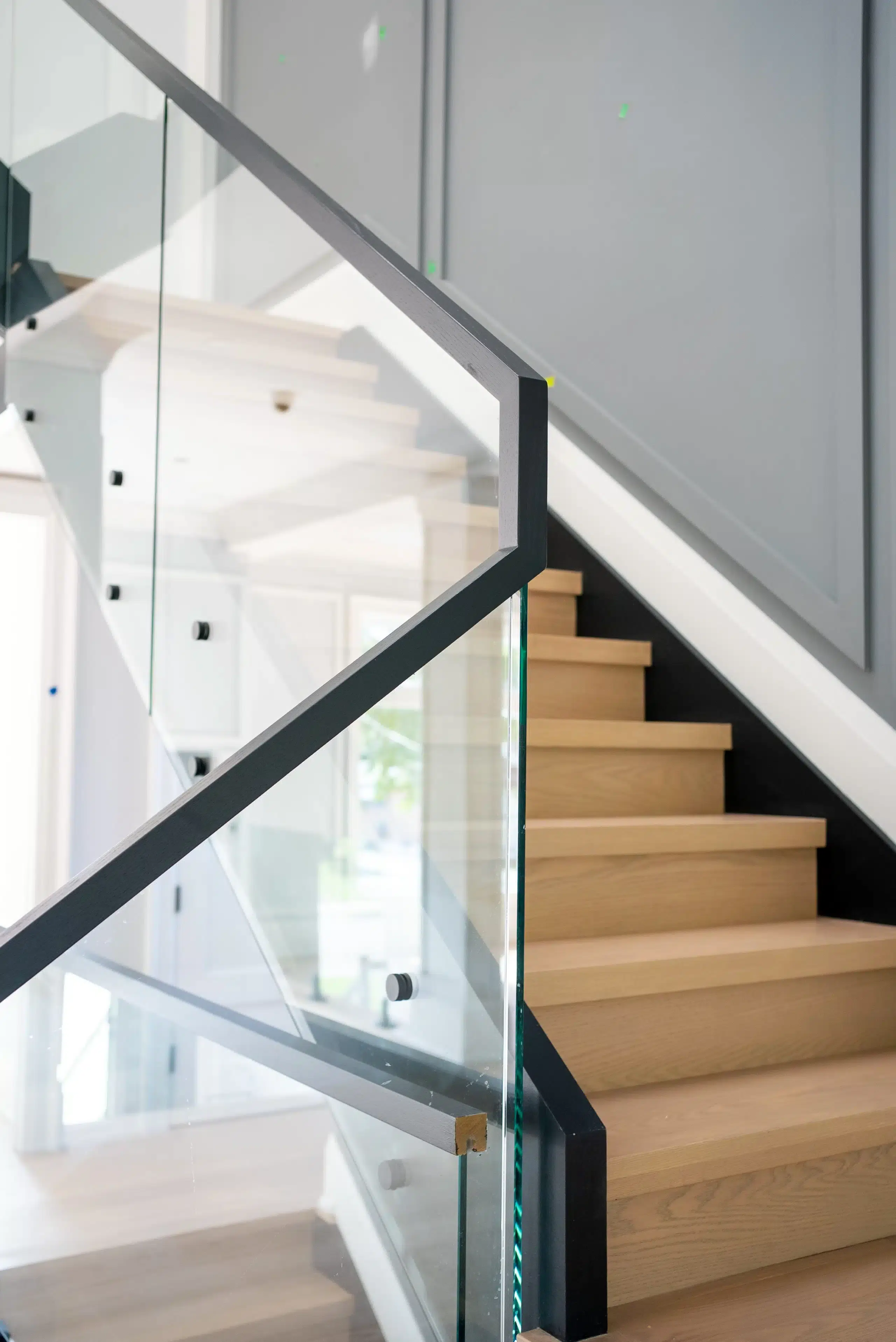 Glass Railing with Black Handrail Close up of a glass railing with a black angled handrail and black standoffs next to white oak stairs