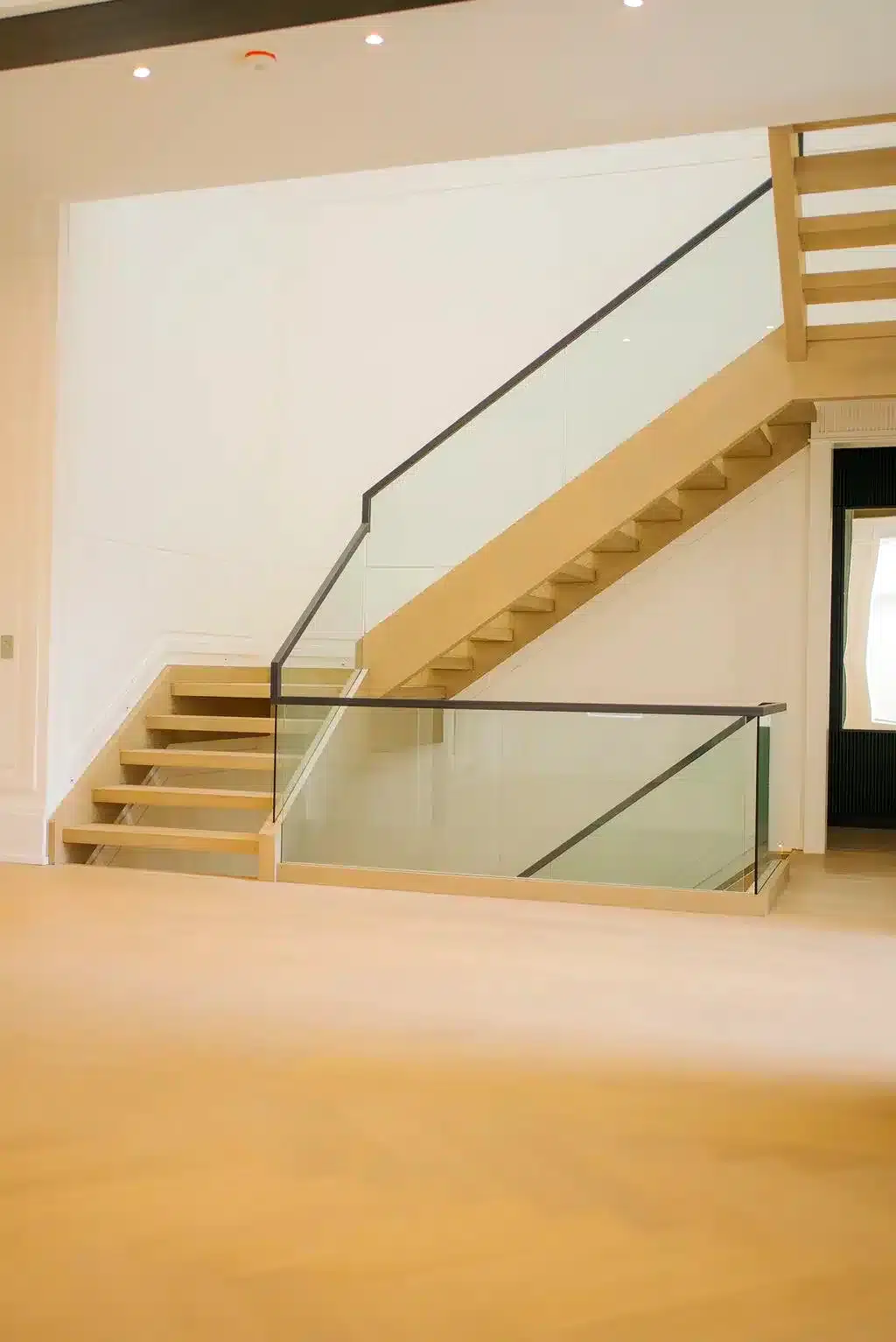 Glass Railing on Light Oak Stairs Frameless glass railing with a slim dark handrail along a light oak open riser staircase and main floor opening