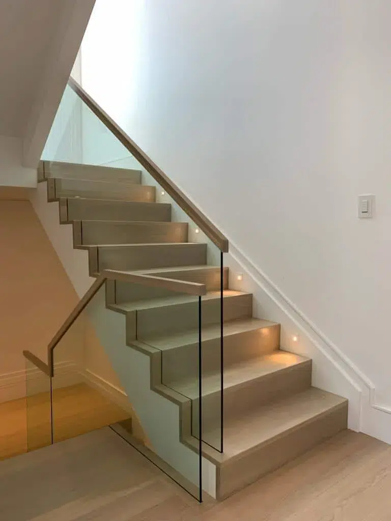 Glass railing with a light wood handrail along a home staircase with light grey treads and recessed step lights
