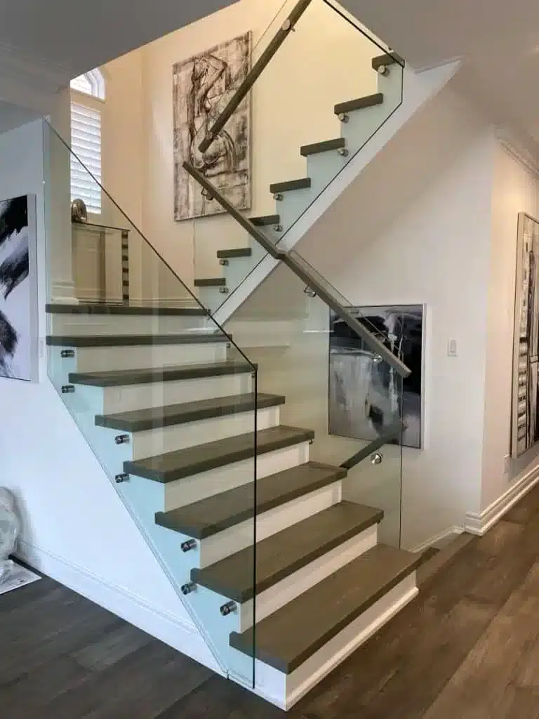 Side-Mounted Frameless Glass Railing Frameless glass railing fixed with stainless steel standoffs and a grey wood handrail on a staircase with grey treads and white risers