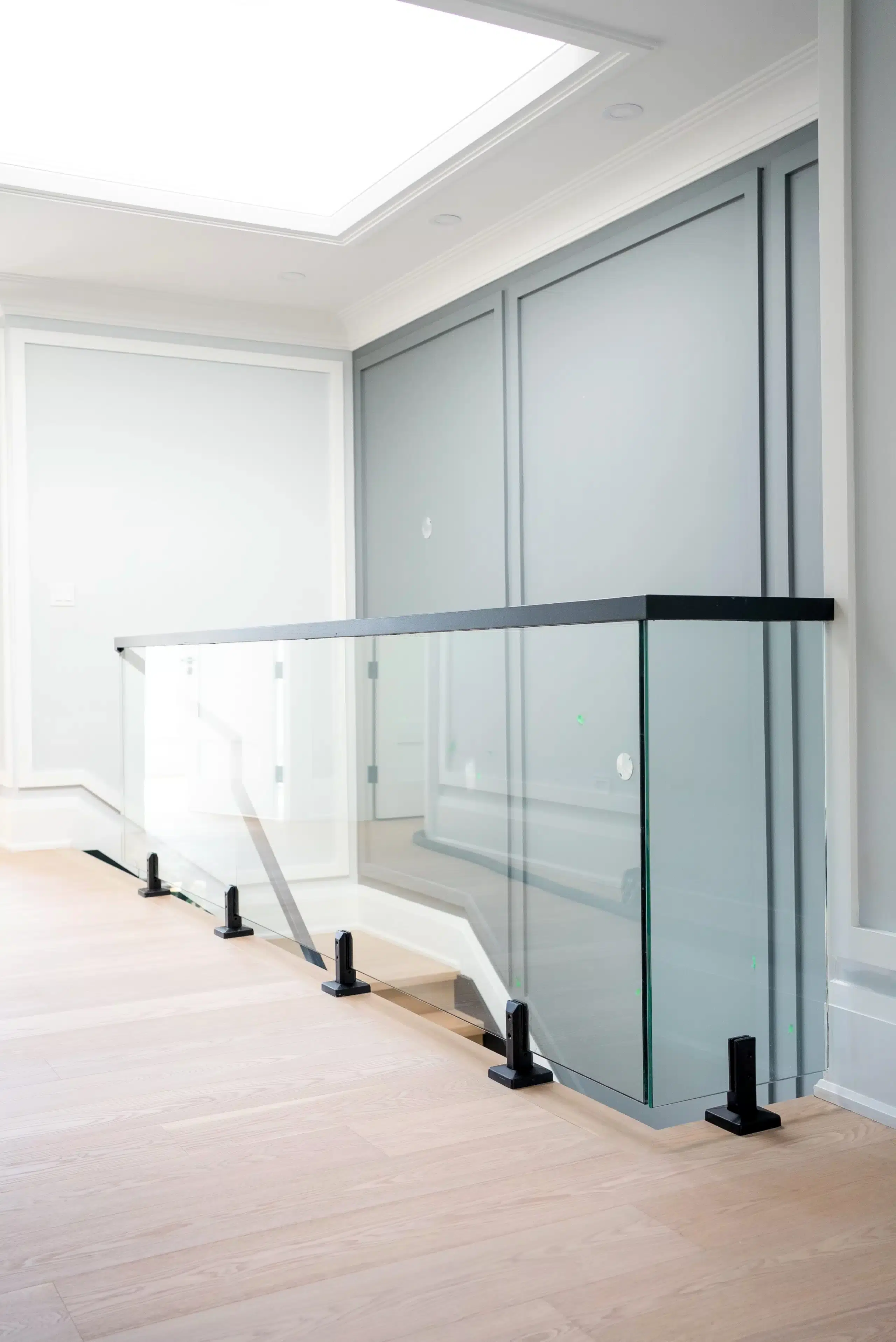 Spigot-Mounted Glass Railing on Landing Spigot mounted glass railing with black base clamps and a black top cap guarding an upper floor stair opening under a skylight